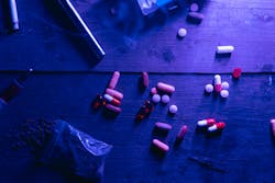 A flat lay of various drugs including pills, capsules, and a syringe on a wooden surface under moody blue lighting.