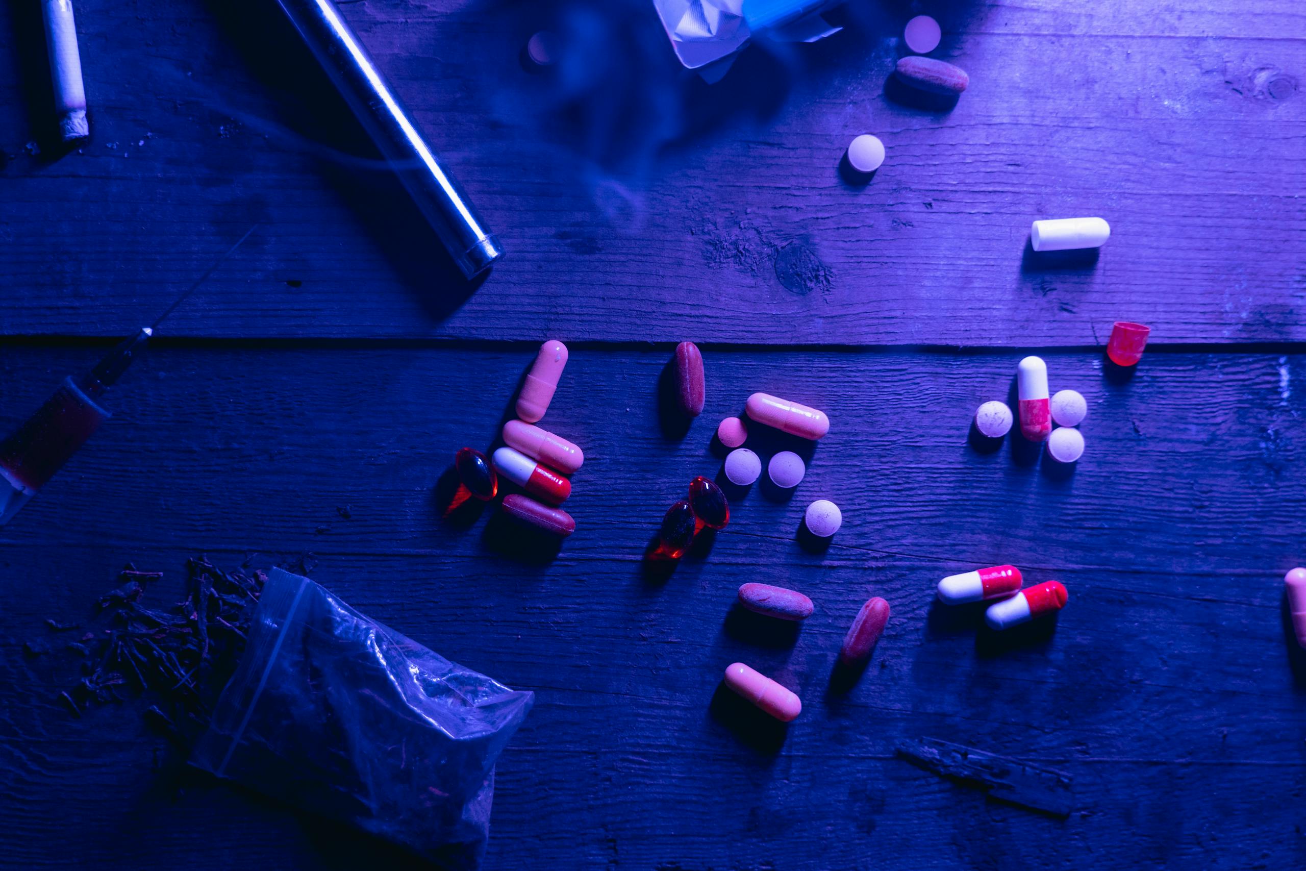 A flat lay of various drugs including pills, capsules, and a syringe on a wooden surface under moody blue lighting.