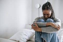 A young woman embraces her knees while sitting on a bed, appearing thoughtful and introspective.