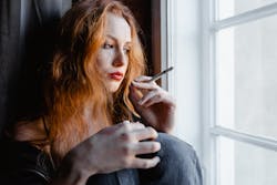 Pensive young woman with red hair holding a cigarette by a window, reflecting indoors.