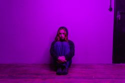 A woman in a hoodie sits alone on the floor, illuminated by purple light indoors.