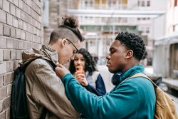 Side view of multiracial classmates bullying student near brick wall and filming video on smartphone
