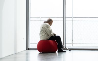 A person sits alone on a red chair in a minimalist interior, gazing outside.